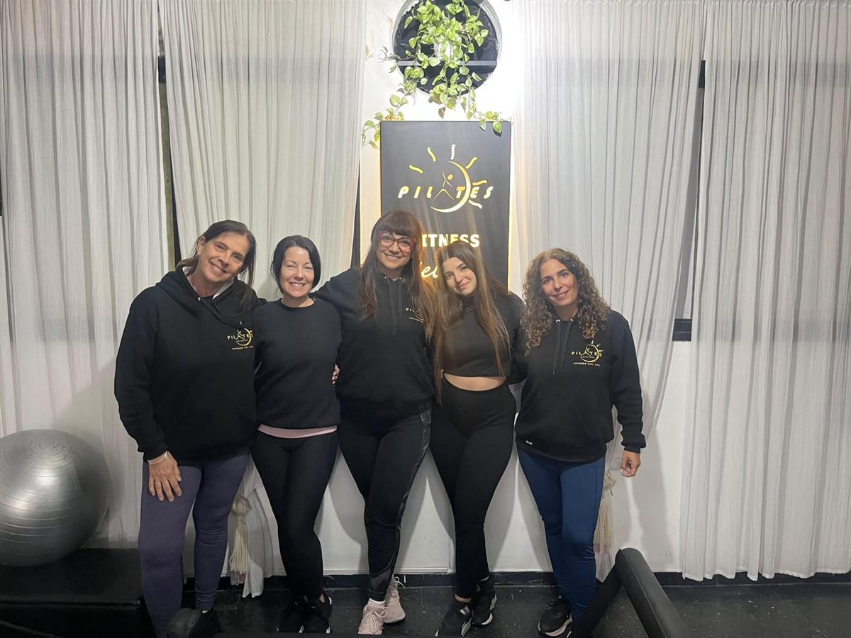 Cinco mujeres con buzos de Fitness del Sol, abrazadas frente al cartel del salón